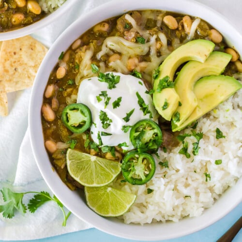 Close up view of Pork Chili Verde in a large bowl. Bowl also contains white rice, jack cheese, sour cream, avocado, cilantro, lime and jalapeños.