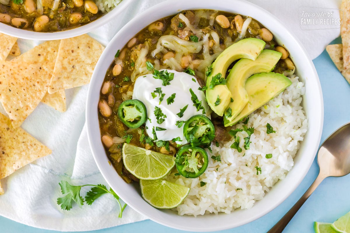 Close up view of Pork Chile Verde in a large bowl. Bowl also contains white rice, jack cheese, sour cream, avocado, cilantro, lime and jalapeños.