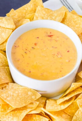 Round serving dip tray with the center bowl containing Nacho Cheese Dip and tortilla chips in the outer bowl surrounding the Nacho Cheese Dip. Salsa and Guacamole bowls on the side.