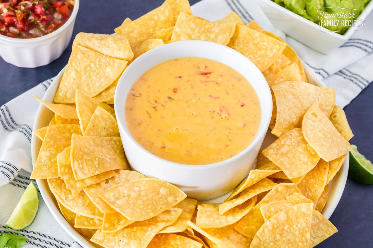 Round serving dip tray with the center bowl containing Nacho Cheese Dip and tortilla chips in the outer bowl surrounding the Nacho Cheese Dip. Salsa and Guacamole bowls on the side.