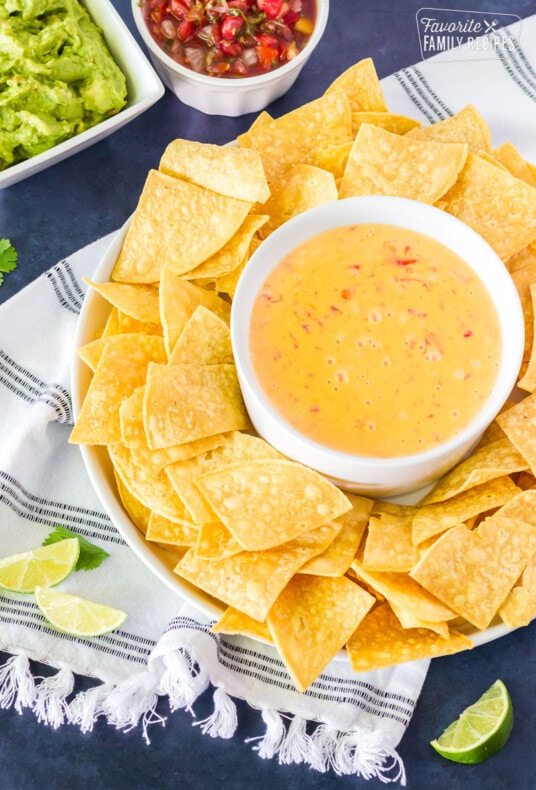 Warmed Nacho Cheese Dip in a bowl surrounded by tortilla chips. Sliced lime wedges, guacamole and salsa on the side.