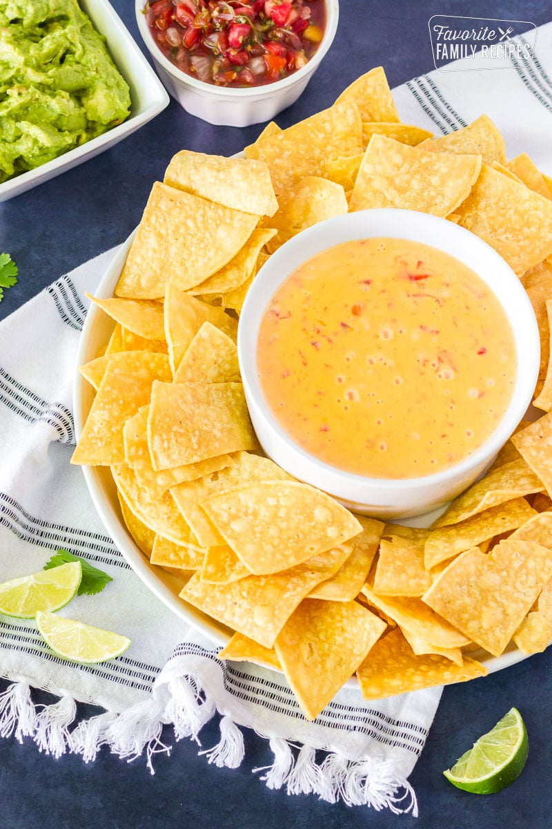 Warmed Nacho Cheese Dip in a bowl surrounded by tortilla chips. Sliced lime wedges, guacamole and salsa on the side.