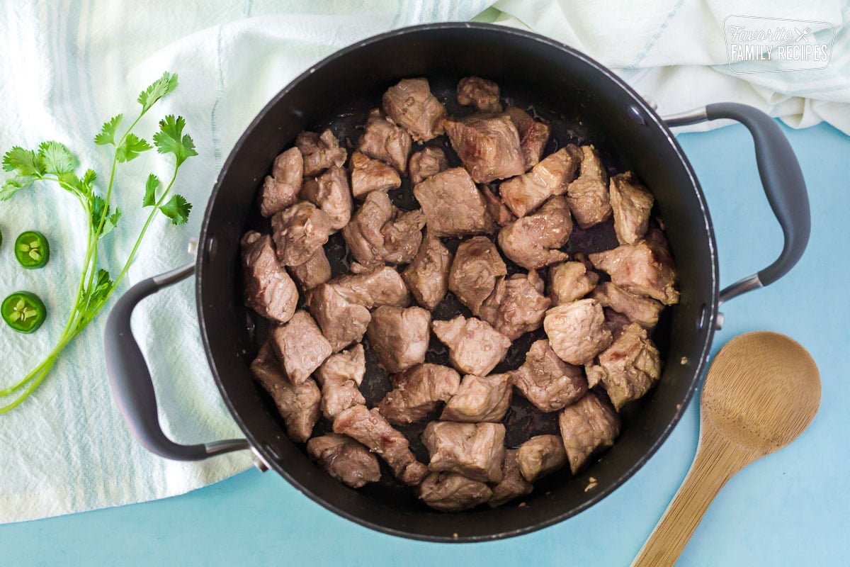 Large pot with browned pork shoulder and wooden spoon on the side to make Pork Chili Verde.