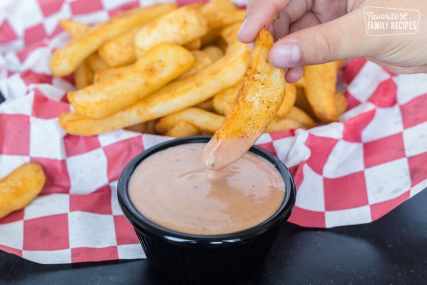 A basket of fries with one French fry being dipped into sauce.