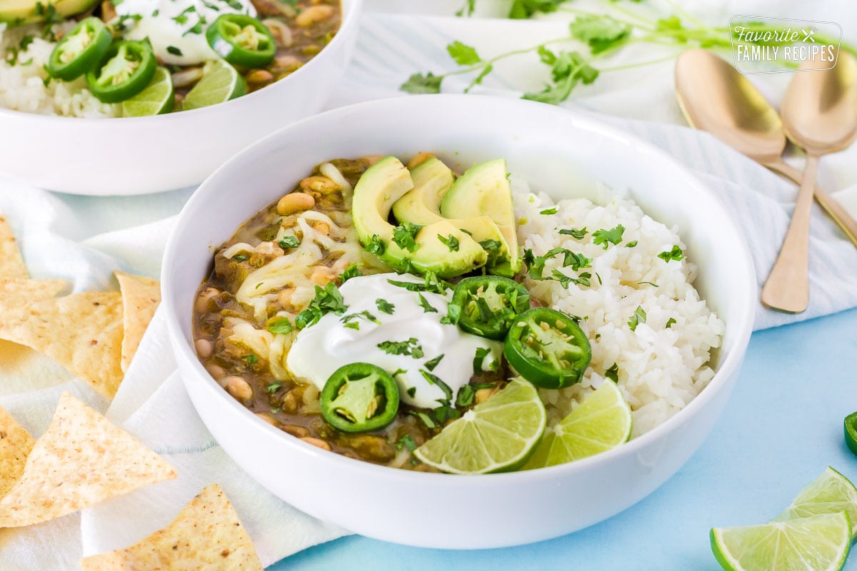 Side view of Pork Chile Verde in a large bowl. Garnished with avocado, sour cream, jalapeños, limes, cilantro and jack cheese.