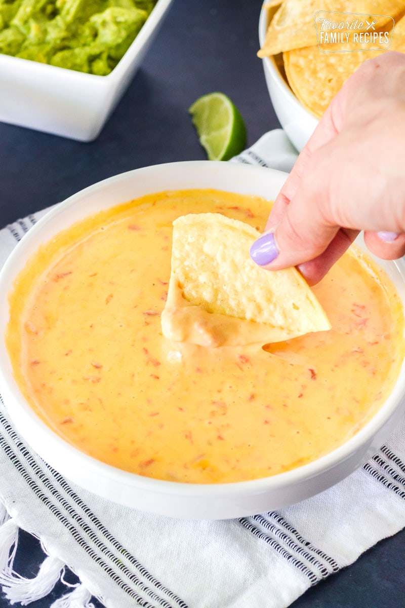 Hand dipping a tortilla chip into a warm bowl of Nacho Cheese Dip. Guacamole on the side.