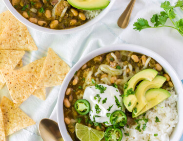 Two bowls of Pork Chili Verde served with white rice, jack cheese, jalapeños, sour cream limes and avocado.