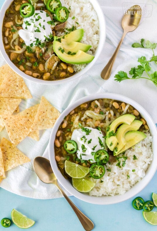 Two bowls of Pork Chili Verde served with white rice, jack cheese, jalapeños, sour cream limes and avocado.