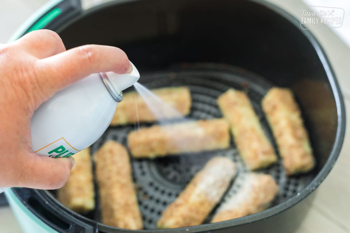 Cooking spray being sprayed on zucchini spears in an air fryer.