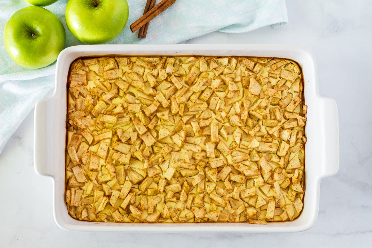 Baked Salted Caramel Apple Cheesecake Bars in a dish. Green apples and cinnamon sticks on the side.