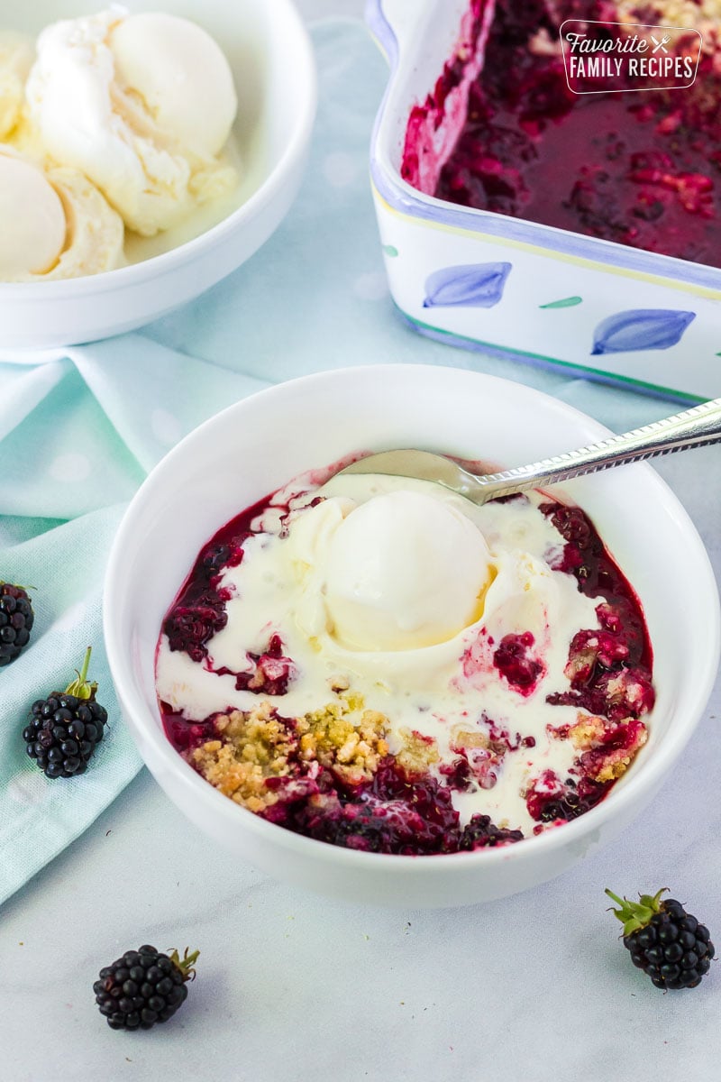Bowl of Easy Blackberry Cobbler with a scoop of melting vanilla ice cream and a spoon. Bowl of ice cream and pan of blackberry cobbler in the background.