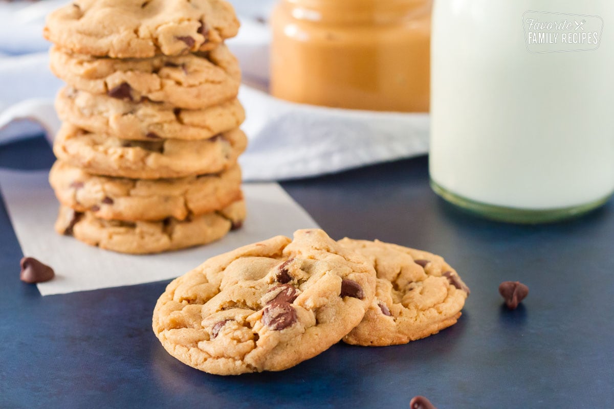 Two Peanut Butter Chocolate Chip Cookies in front of a stack of cookies. Glass of milk and peanut butter on the side.