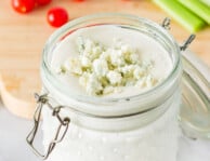 Close up view of Blue Cheese Dressing in a jar topped with crumbles. Tomatoes and celery on the side.