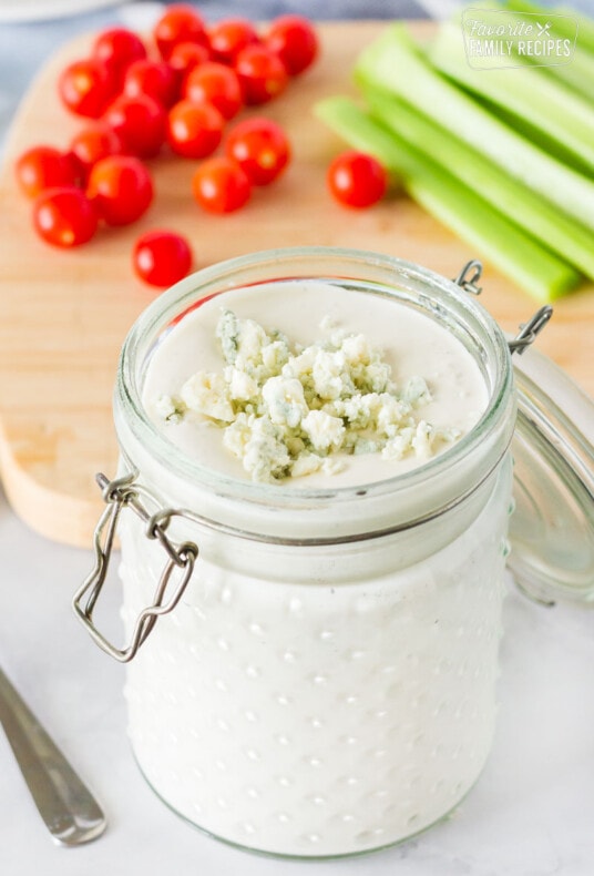 Close up view of Blue Cheese Dressing in a jar topped with crumbles. Tomatoes and celery on the side.