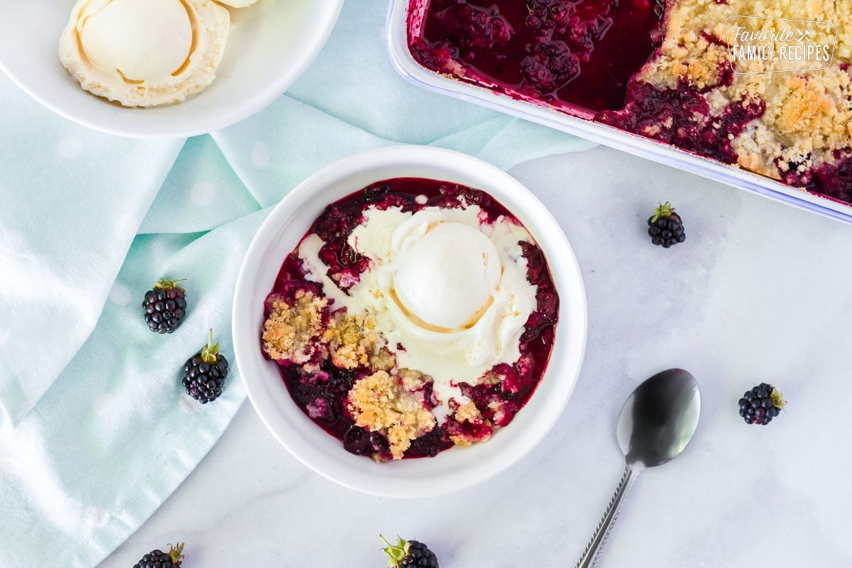 Top view of a bowl of Easy Blackberry Cobbler with melted vanilla ice cream. Separate bowl of vanilla ice cream scoops and pan of Blackberry Cobbler.