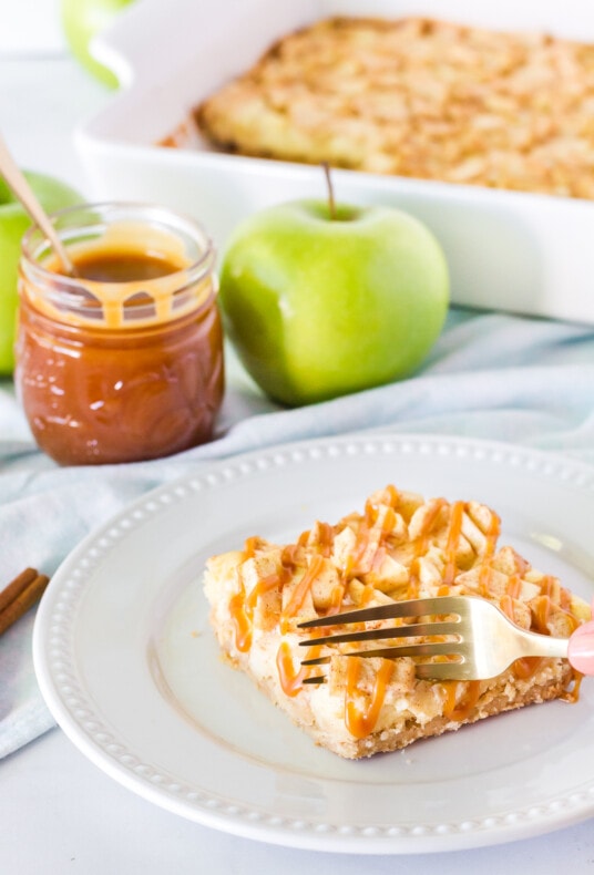 Fork cutting into a slice of Salted Caramel Apple Cheesecake Bar. Caramel, apples and dish in the background.