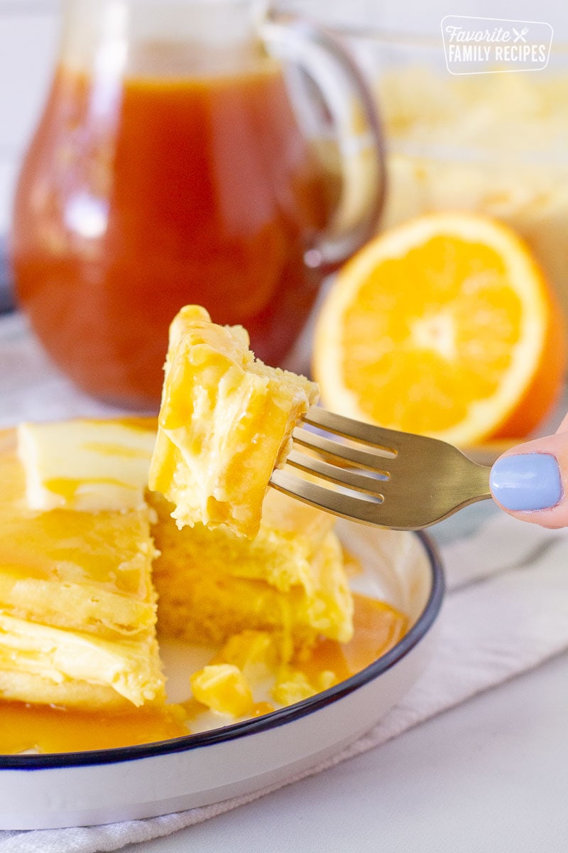 Bite of an orange pancake on a golden fork. Plate of pancakes and orange syrup in the background.