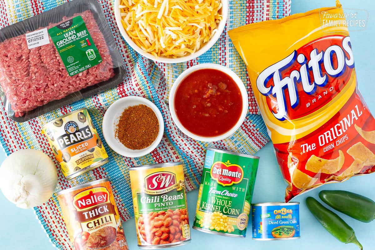 Ingredients to make Frito Chili Pie including: bag of Fritos, shredded cheese, ground beef, salsa, taco seasoning, onion, can of green chilies, kernel corn, chili pinto beans, chili, sliced black olives and fresh jalapeños as a garnish.