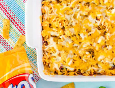 Frito Chili Pie in a serving dish with a bag of Fritos on the side. Frito chips and sliced jalapeños are around the dish.