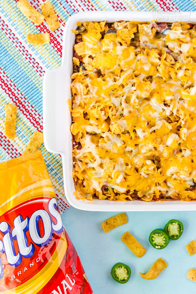 Frito Chili Pie in a serving dish with a bag of Fritos on the side. Frito chips and sliced jalapeños are around the dish.