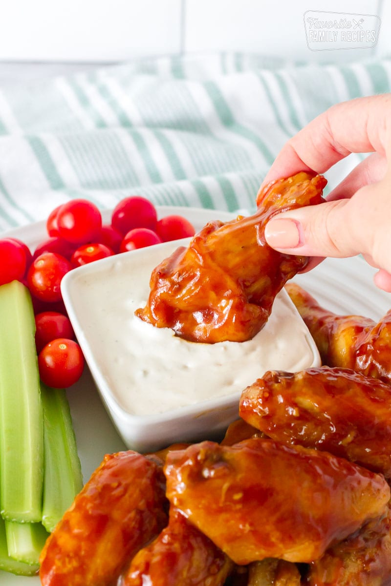 Hand Dipping Hot Wing into a small bowl of Blue Cheese Dressing. Celery and tomatoes on the side.