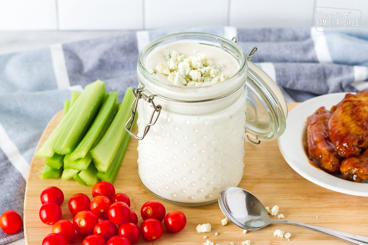 Blue Cheese Dressing in a jar topped with crumbles. Sides of sliced celery, hot wings and tomatoes.