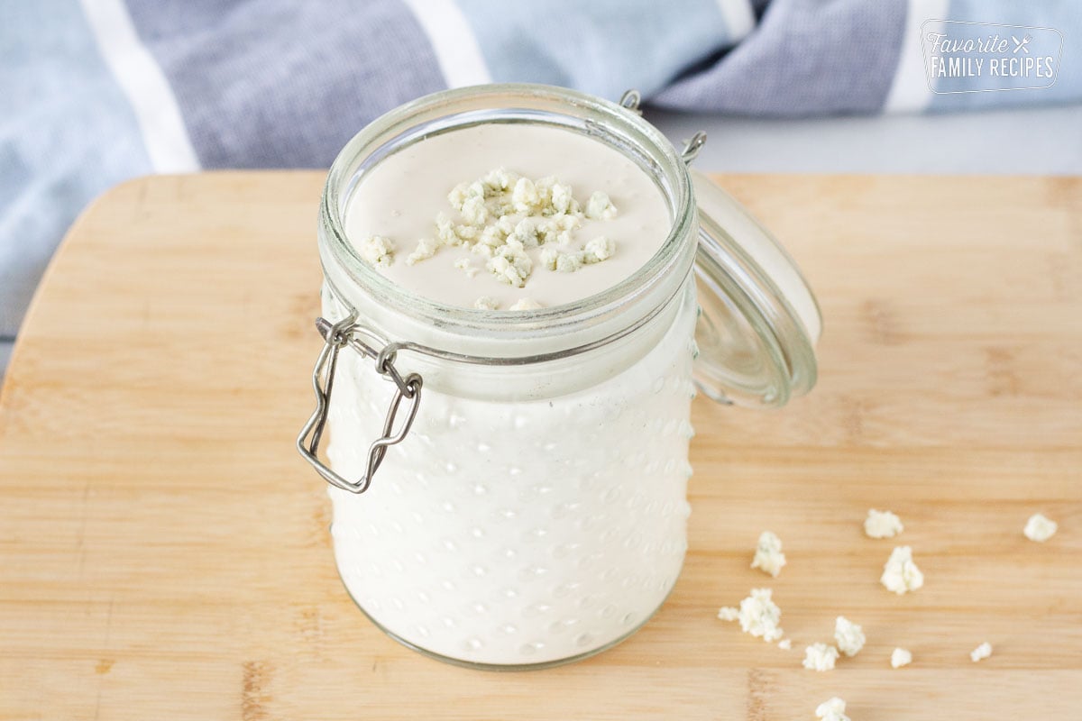 Jar of Blue Cheese Dressing topped with blue cheese crumbles on a cutting board.