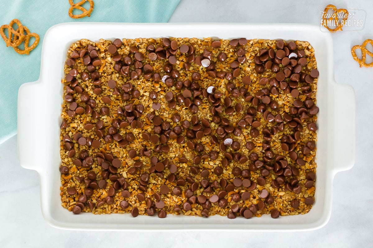 Baking dish with baked pretzel crust topped with melted chocolate chips for Pretzel Crust Cheesecake Brownies.