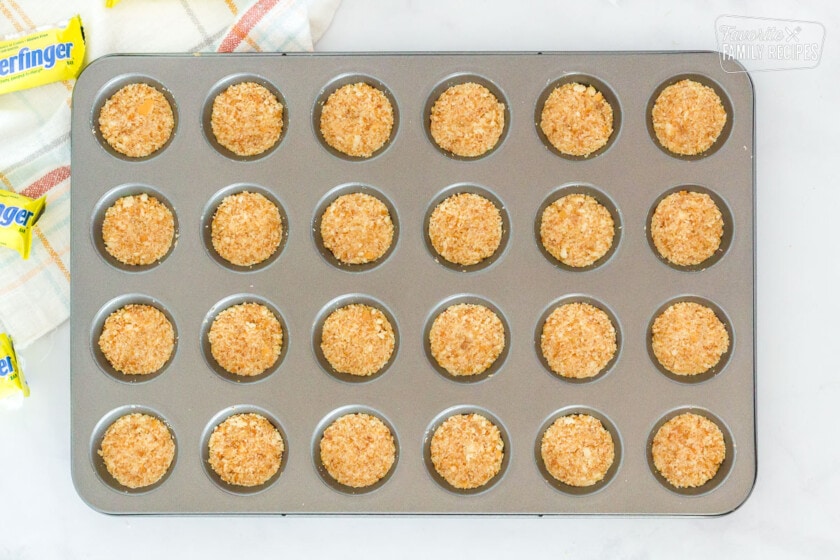 Mini muffin pan with pressed crust mixture for Butterfinger Bites. Butterfinger candy bars on the side.