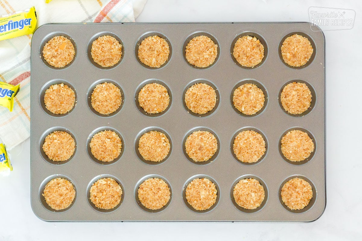 Mini muffin pan with pressed crust mixture for Butterfinger Bites. Butterfinger candy bars on the side.