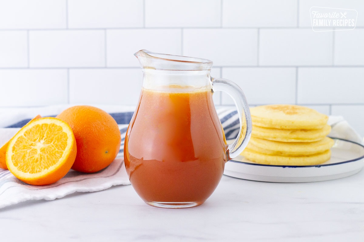 Orange syrup in a pitcher for Orange Pancakes. Plate of Orange Pancakes and fresh oranges on the side.