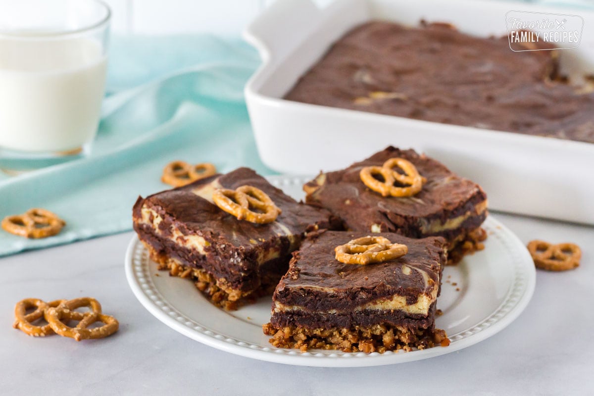 Plate of three Pretzel Crust Cheesecake Brownies with a glass of milk. Baking dish of Pretzel Crust Cheesecake Brownies in the background.