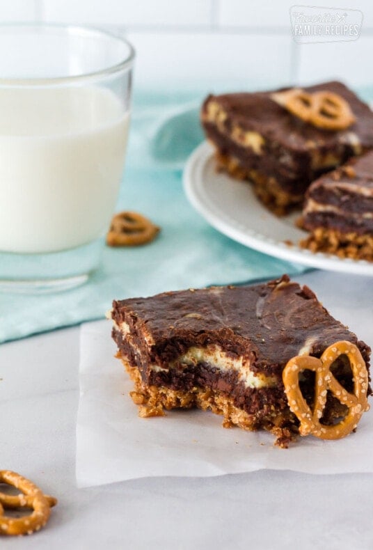 Pretzel Crust Cheesecake Brownie with a bite taken out. Single pretzel on the side and glass of milk.