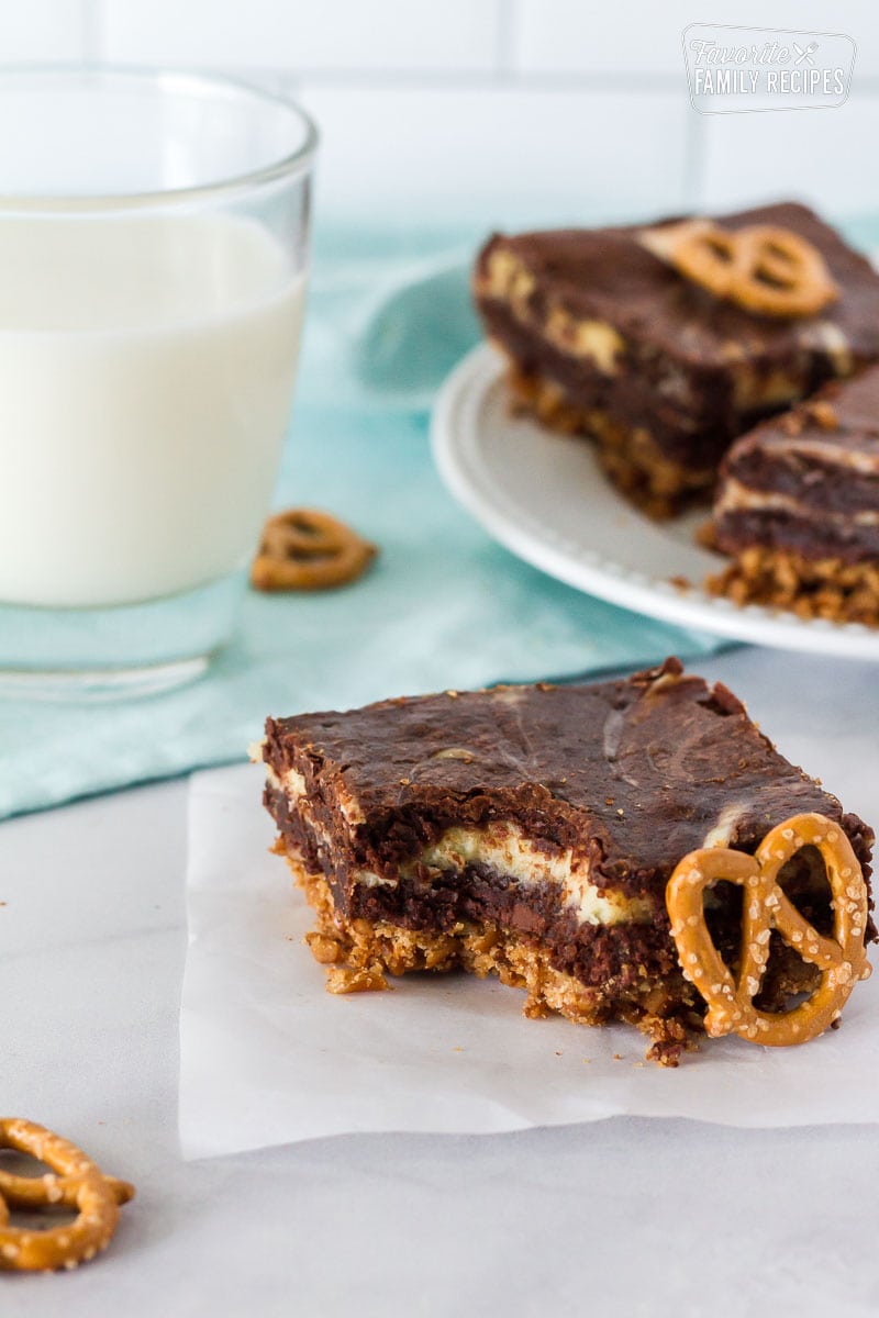 Pretzel Crust Cheesecake Brownie with a bite taken out. Single pretzel on the side and glass of milk.