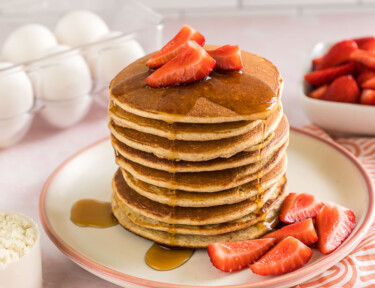A stack of protein pancakes with strawberries on top and maple syrup dripping down the side