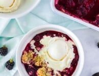 Bowl of Easy Blackberry Cobbler with a scoop of vanilla ice cream melting on top. Side bowl of ice cream and pan of cobbler.