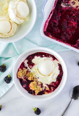 Bowl of Easy Blackberry Cobbler with a scoop of vanilla ice cream melting on top. Side bowl of ice cream and pan of cobbler.