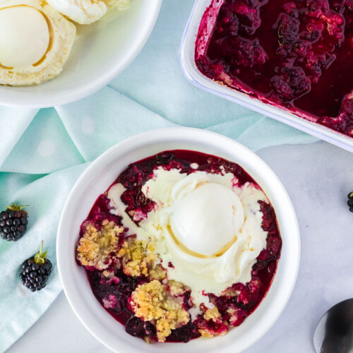 Bowl of Easy Blackberry Cobbler with a scoop of vanilla ice cream melting on top. Side bowl of ice cream and pan of cobbler.