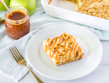 Caramel Apple Cheesecake Bar on a plate with a fork and extra caramel on the side. Dish of Apple Cheesecake Bars and green apples in the background.
