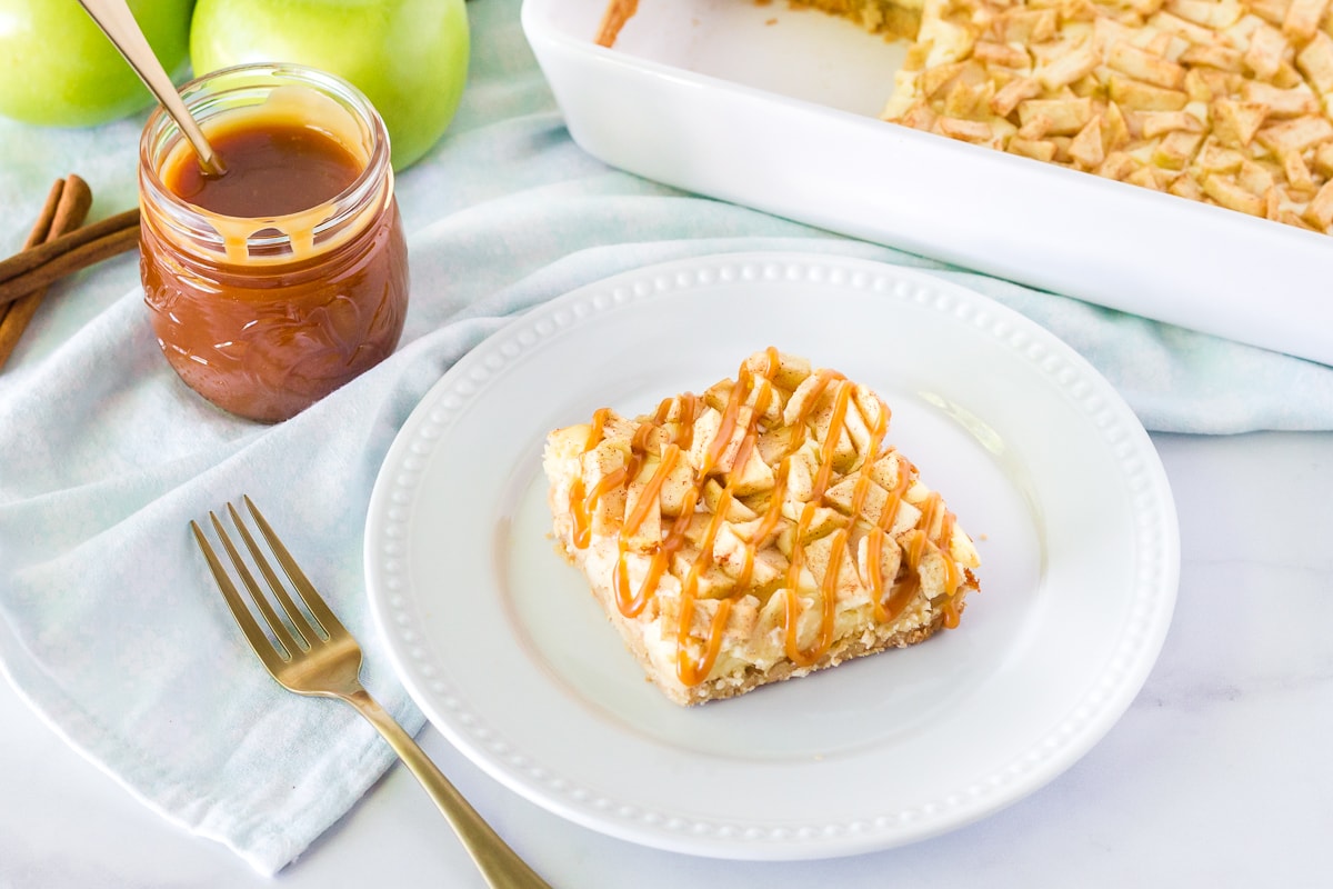 Caramel Apple Cheesecake Bar on a plate with a fork and extra caramel on the side. Dish of Apple Cheesecake Bars and green apples in the background.