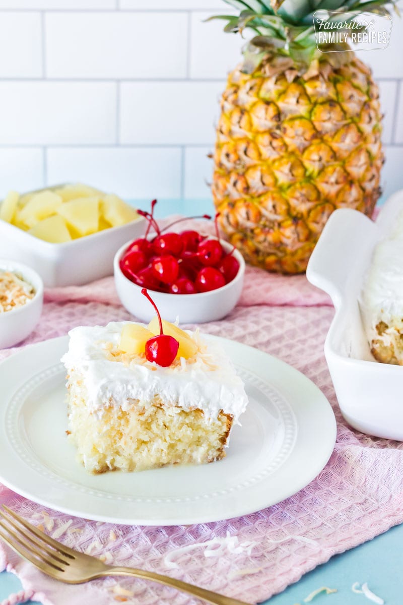 Slice of Pina Colada Cake on a plate. Fresh pineapple, cherries, toasted coconut and pineapple slices on the side.