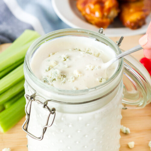 Spoon scooping up Blue Cheese Dressing from a jar. Side of celery, tomatoes and hot wings.