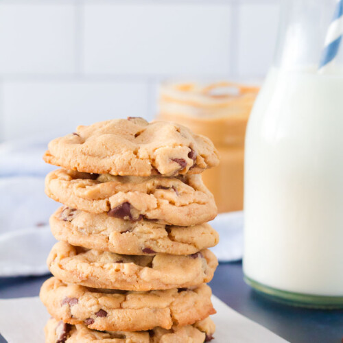 Tall stack of six Peanut Butter Chocolate Chip Cookies on a square of parchment paper. Glass of milk served on the side.
