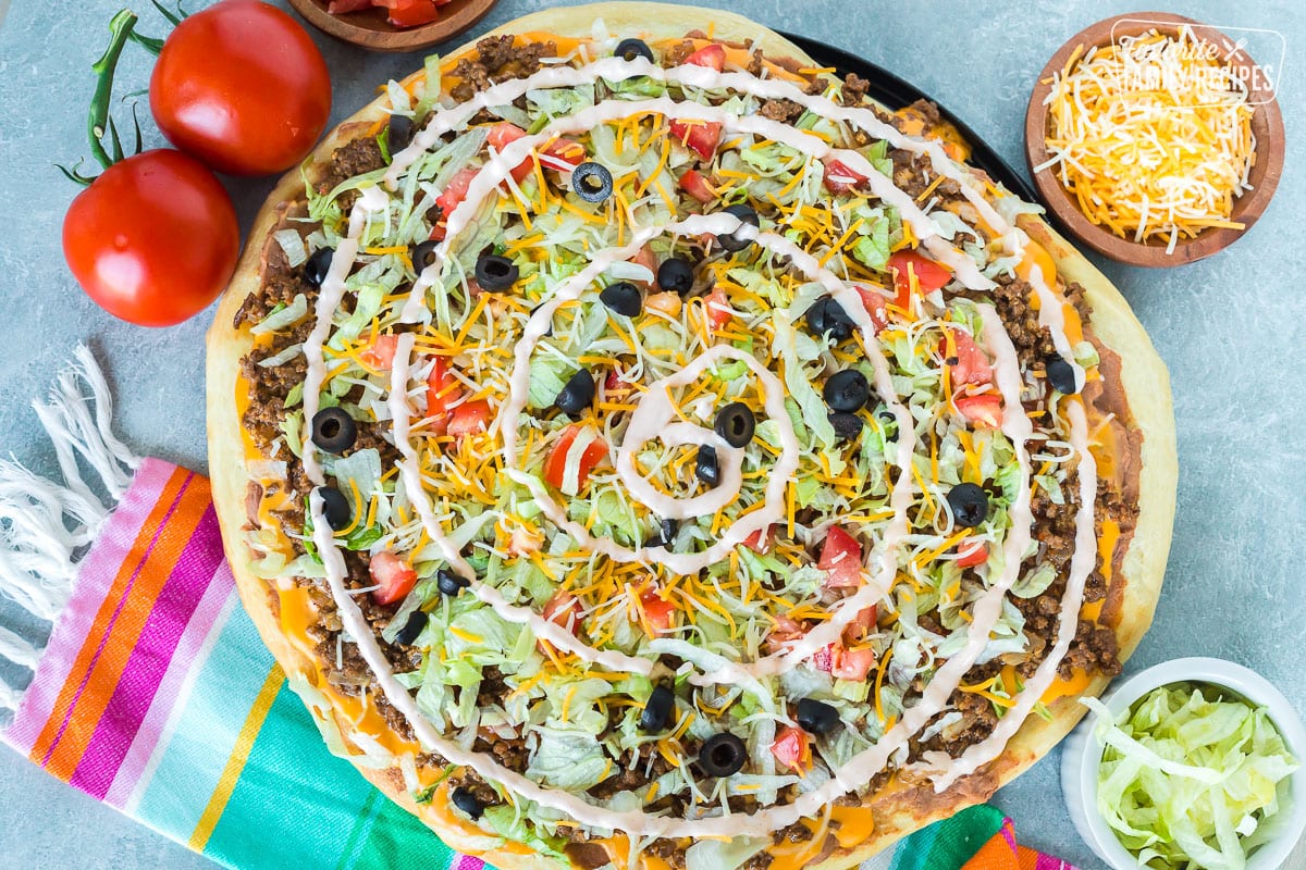 A top view of a taco pizza next to tomatoes, shredded cheese, and shredded lettuce.