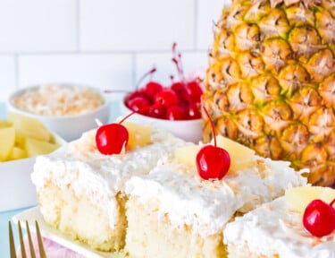 Three slices of Pina Colada Cake in a row. Fresh pineapple, cherries and toasted coconut on the side.