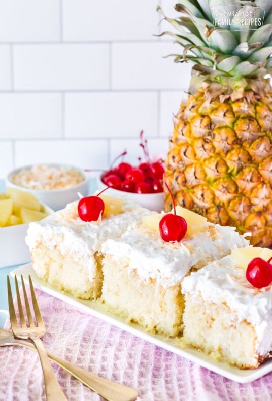 Three slices of Pina Colada Cake in a row. Fresh pineapple, cherries and toasted coconut on the side.