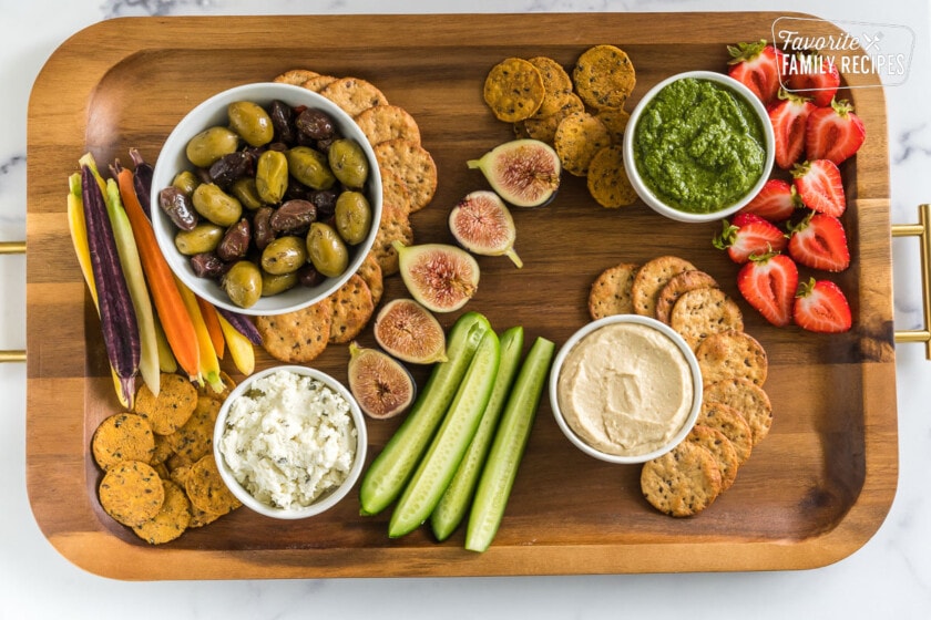 A wooden serving plate with 4 white bowls and various crackers, nuts, fruit, and vegetables