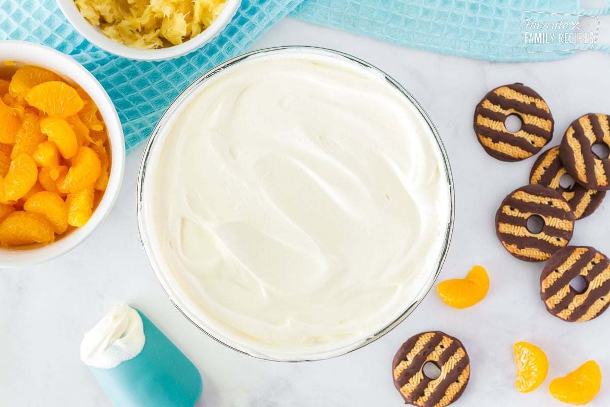 Bowl of whipped topping mixed with pudding. Bowls of mandarin oranges, crushed pineapple, fudge stripe cookies and mandarin oranges on the side.