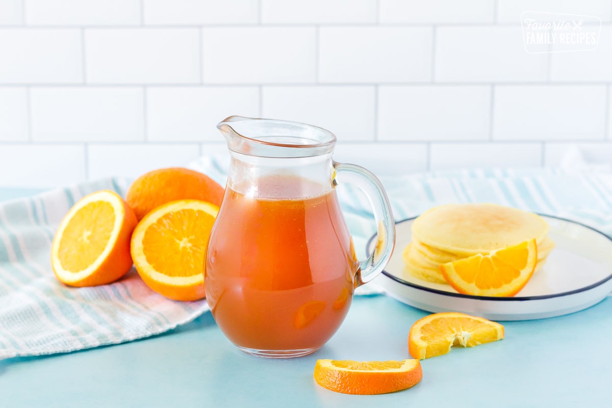 Orange Syrup in a pitcher with a plate of pancakes and orange slices.
