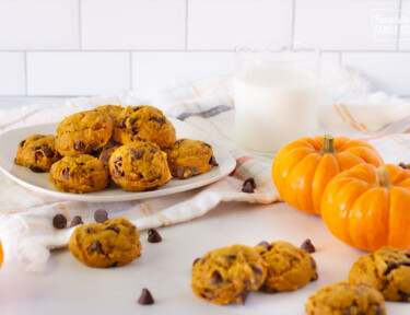 Fresh Pumpkin Chocolate Chip Cookies on a plate with a side of milk. Mini pumpkins and extra cookie decorate the table.